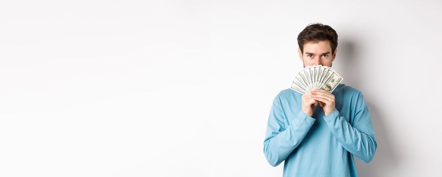 Finance concept. Smiling young man covering face behind money, showing dollars and looking happy, standing over white background.