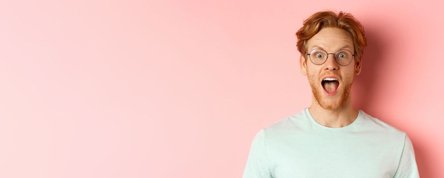Close up of surprised redhead man raising eyebrows and scream with joy, checking out cool promo, looking amazed at camera, standing over pink background.