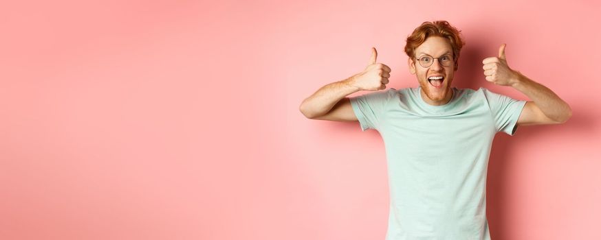 Amazed redhead guy, scream with joy and looking impressed and excited, showing thumbs up in approval, praise awesome promo offer, standing over pink background.