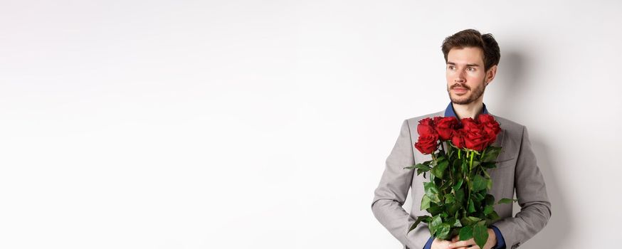 Handsome boyfriend in suit going on romantic date, holding bouquet of red roses and looking left thoughtful, standing over white background.