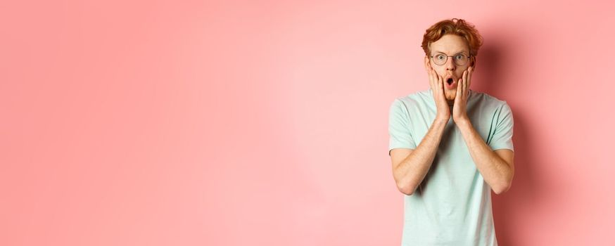 Shocked redhead man gossiping, staring impressed at camera and touching face, standing in glasses against pink background.
