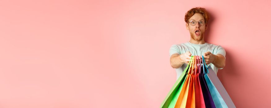 Surprised redhead man stretch out hands with shopping bags, give you gifts, standing over pink background.
