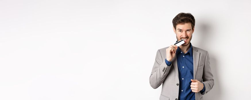 Successful young businessman in suit biting his credit card and looking sassy, standing on white background.