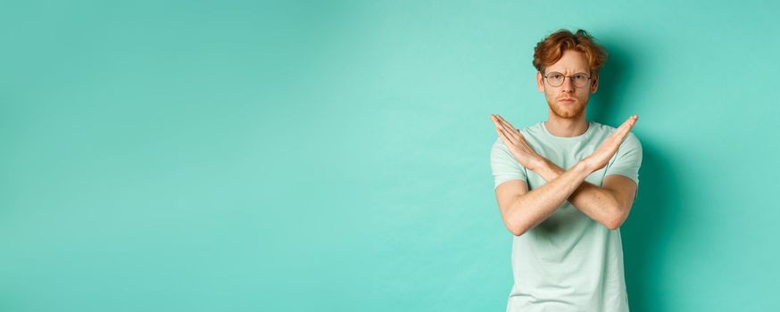 Serious and confident redhead man in t-shirt and glasses saying no, showing cross gesture to stop you, refucing or declining something, standing over turquoise background.