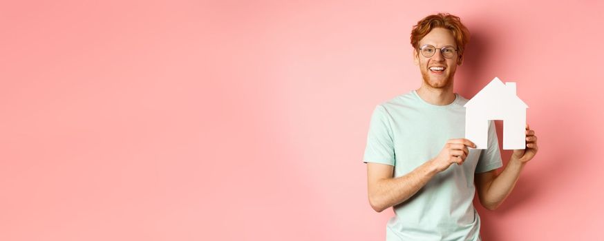 Real estate. Handsome redhead man in t-shirt and glasses, showing paper house cutout and smiling, standing over pink background.