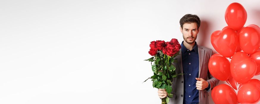 Valentines day. Handsome and confident man going on date in suit, holding bouquet of red roses and heart balloons, standing with gifts for lover on white background.