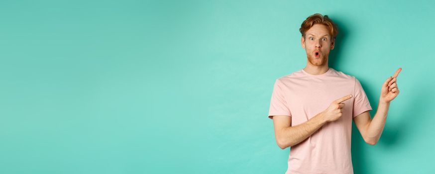 Handsome caucasian man with red hair, gasping in awe, pointing fingers at upper right corner, checking out promotion deal, standing over turquoise background.