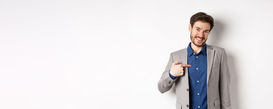 Modest smiling man in suit pointing at himself with cute face, self-promoting, standing on white background.