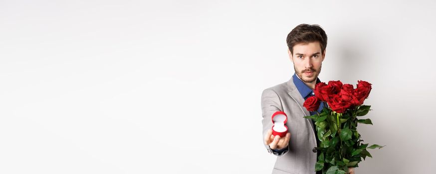 Handsome young man making a marriage proposal, stretch out hand with engagement ring and holding red roses, asking to marry him, looking confident at lover, white background.