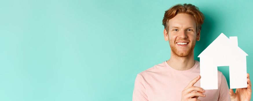 Real estate concept. Close up of redhead bearded man in t-shirt showing paper house cutout, smiling happy at camera, standing over turquoise background.