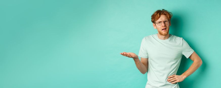 Confused redhead man trying to understand something, squinting clueless and stretch out one hand, looking puzzled, standing over mint background. Copy space