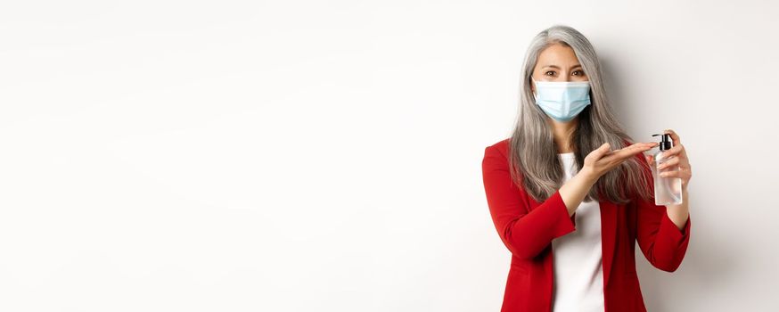 Covid, pandemic and business concept. Asian female manager in medical mask using hand sanitizer and smiling at camera, standing with antiseptic over white background.