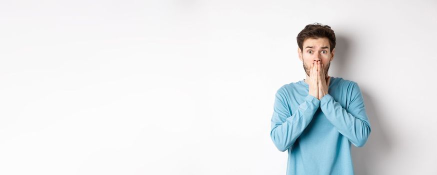 Shocked and worried young man gasping and covering mouth, look with panic at camera, standing anxious against white background.