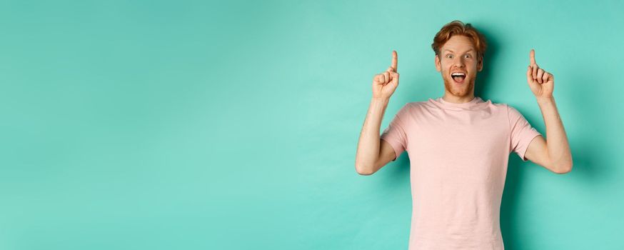 Cheerful redhead man in t-shirt pointing fingers up, staring in awe at camera and showing advertisement, standing over turquoise background.