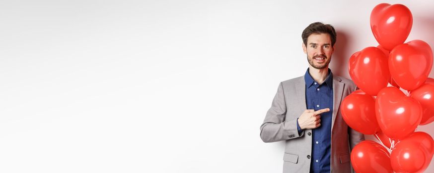 Lovers day. Handsome smiling guy in suit making surprise gift on Valentines date, pointing at romantic heart balloons and looking happy, standing over white background.