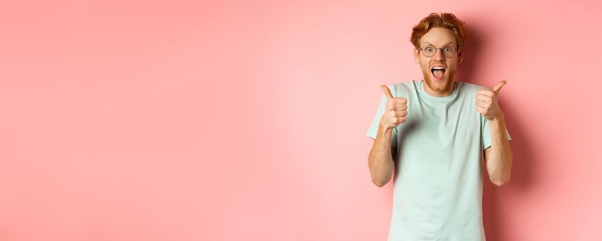 Amazed redhead guy looking impressed and excited, showing thumbs up in approval, praise awesome promo offer, standing over pink background.
