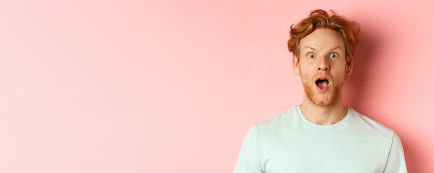 Face of surprised redhead man reacting to cool promo offer, raising eyebrows and gasping, staring in awe at camera, standing over pink background.