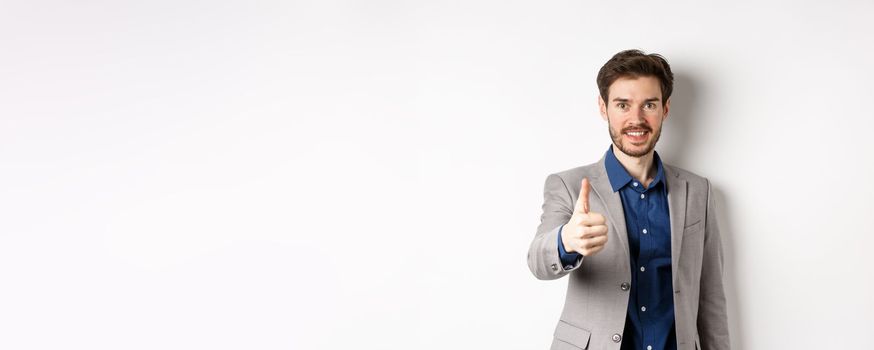 Image of handsome businessman in suit showing thumb up and smiling, standing on white background.