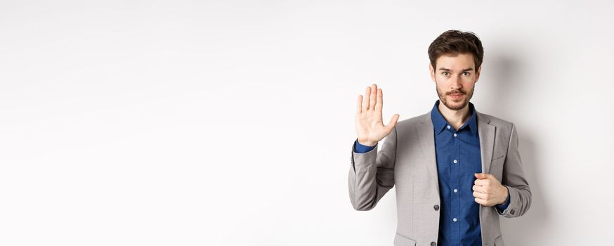 Businessman in formal suit raising hand up to say hello, contactless greeting, smiling friendly, standing on white background.