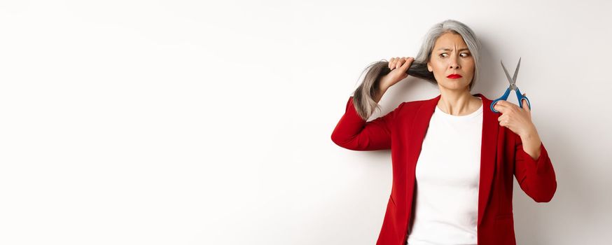 Indecisive asian woman holding scissors and looking doubtful, thinking to cut hair, changing haircut, standing over white background.