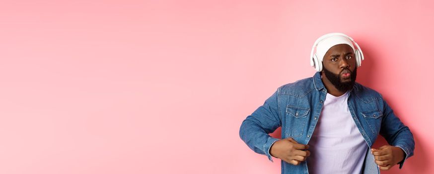 Cool and sassy Black guy dancing, listening to music in headphones, looking confident, standing over pink background. Copy space