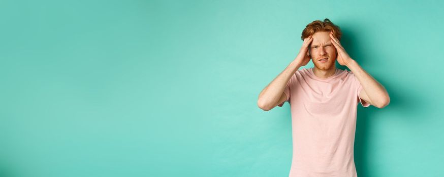 Young redhead man touching head and looking dizzy, feeling headache or migraine, standing in t-shirt over mint background.