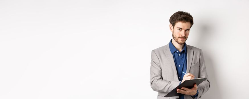 Successful businessman in grey suit taking notes, working in office clothing, holding clipboard and looking at camera, white background.