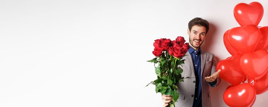 Romantic guy in suit saying happy Valentines day, giving red roses to you and pointing hand, standing with heart balloon gift over white background.
