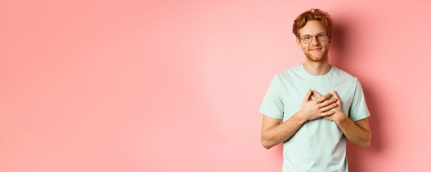 Attractive young man with ginger hair, holding hands on heart and smiling grateful, saying thank you, express gratitude, standing over pink background.