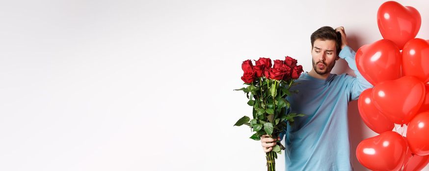 Valentines day romance. Confused boyfriend scratch head and looking at bouquet of red roses for his date. Man with flowers and balloons feeling indecisive, white background.