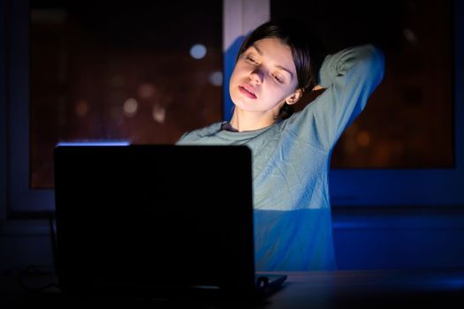 A young girl works while sitting at a laptop at night at home online, a woman is looking for information, texting, chatting using device via internet.