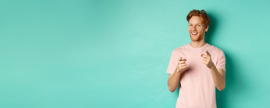 Funny redhead guy congratulating you, pointing fingers at camera and smiling, making choice, standing in t-shirt over mint background.