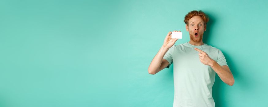 Shopping concept. Handsome redhead man in t-shirt showing plastic credit card and smiling, standing over turquoise background.