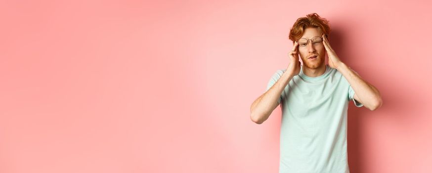 Portrait of redhead man in crooked glasses touching head and feeling dizzy or nauseous, having hangover or headache, standing over pink background.