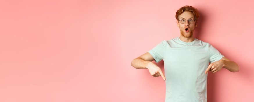 Image of amazed young man with red hair and beard, wearing glasses and t-shirt, pointing fingers down and staring excited at camera, pink background.
