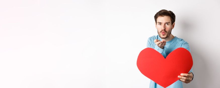 Romantic boyfriend sending air kiss at camera and holding big red Valentines day heart, standing over white background.