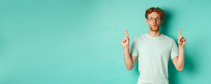 Impressed redhead man in glasses and t-shirt, checking out promo offer, pointing fingers up at copy space, staring at camera amazed, standing over turquoise background.