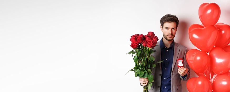 Handsome young man prepare to make proposal, holding engagement ring with bouquet of red roses, making surprise on Valentines day, standing near heart balloons, white background.