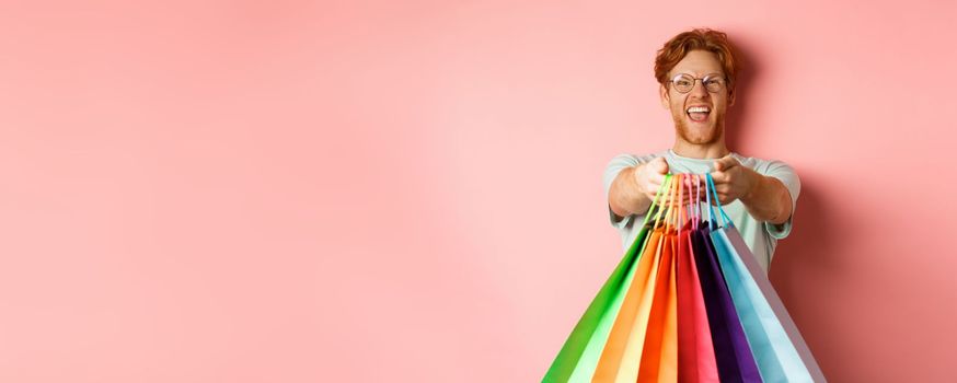 Happy redhead man stretch out hands with shopping bags, give you gifts, standing over pink background.