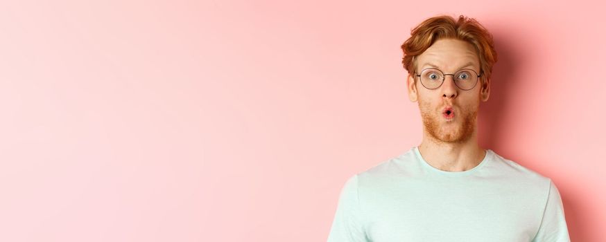 Close up of impressed redhead man in glasses saying wow, raising eyebrows surprised and staring at camera, standing over pink background.
