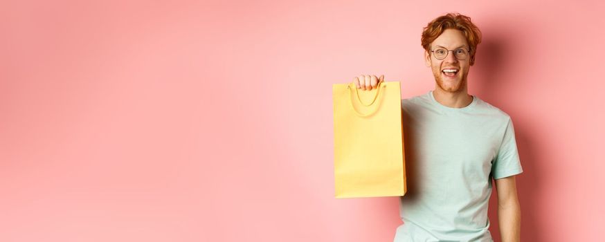 Cheerful redhead man in t-shirt, showing shopping bag and smiling, standing over pink background.
