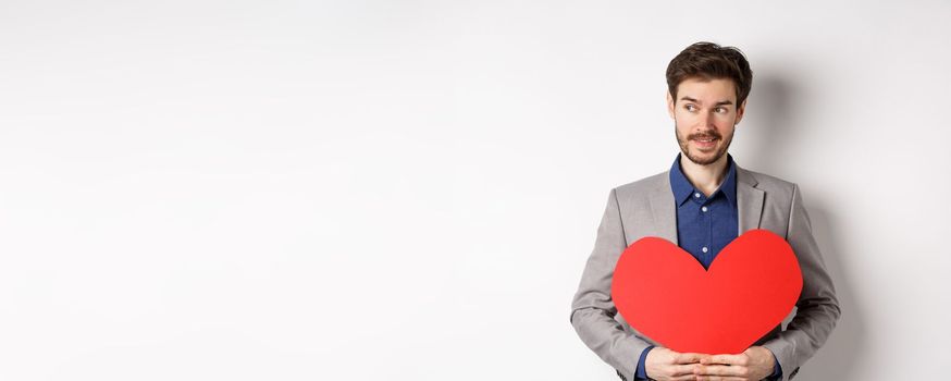 Attractive man in suit looking left and smiling, holding red heart cutout, prepare valentines day surprise for lover, standing over white background.