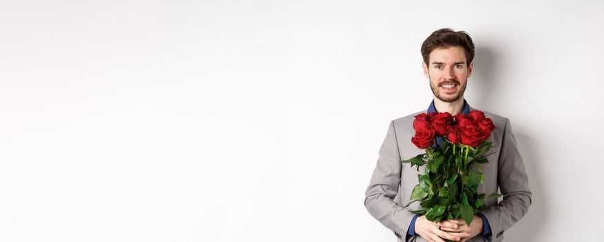 Handsome young man in suit standing with gift on valentines day, holding red roses bouquet and smiling at lover, standing over white background.