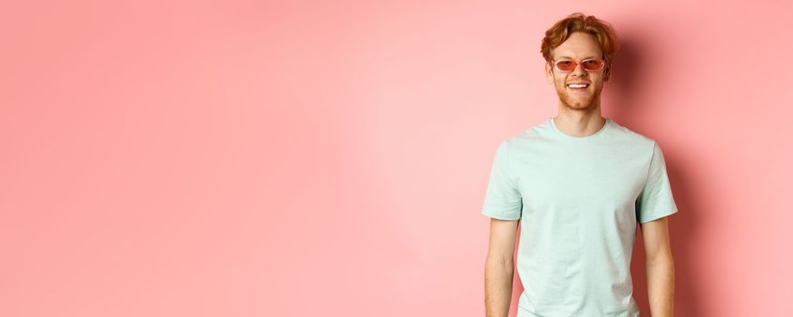 Tourism and vacation concept. Cheerful redhead bearded man in sunglasses and t-shirt, smiling and looking happy at camera standing over pink background.