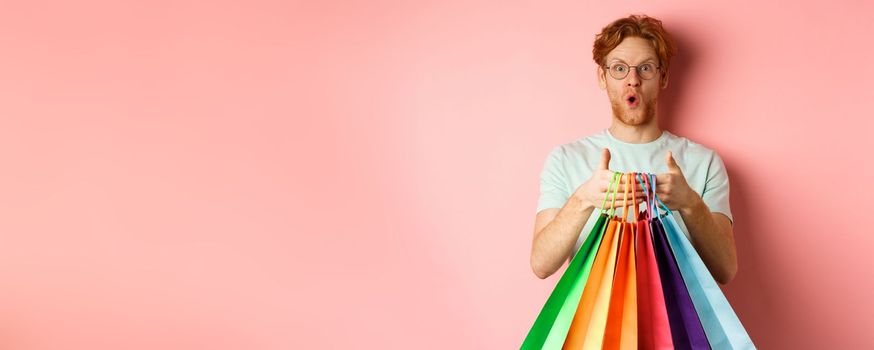 Young handsome man amazed with special discounts, buying gifts, holding shopping bags and standing over pink background.