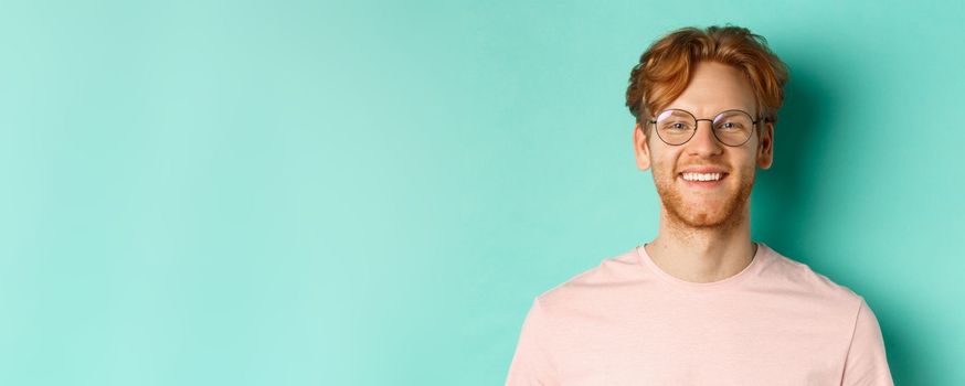 Close up of handsome redhead man in glasses looking at camera, smiling with white teeth, standing against mint background.