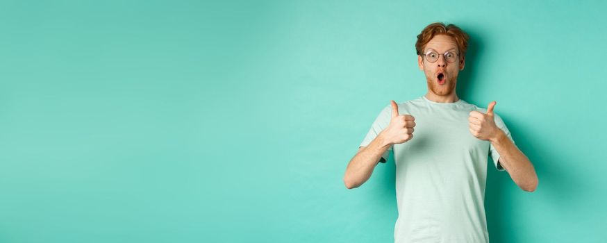 Amazed young man with red hair and beard, wearing glasses with t-shirt, showing thumbs-up and gasping in awe, checking out awesome promo offer, turquoise background.
