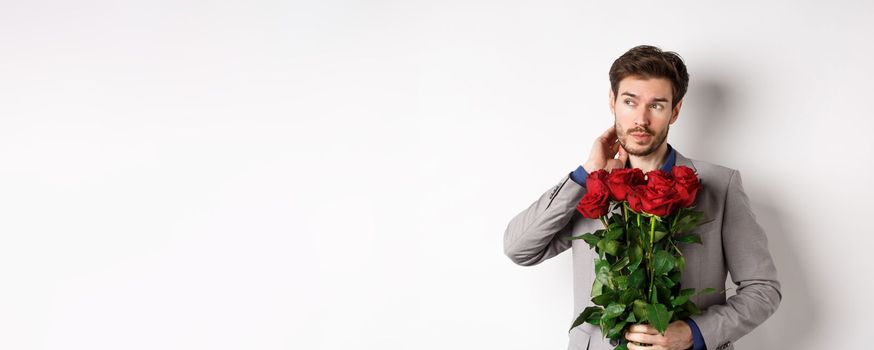 Pensive young man in suit holding bouquet of flowers, waiting for date on Valentines day, standing over white background.