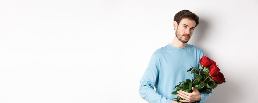 Handsome young man holding beautiful red roses for his lover on Valentines day, looking pensive, standing over white background.