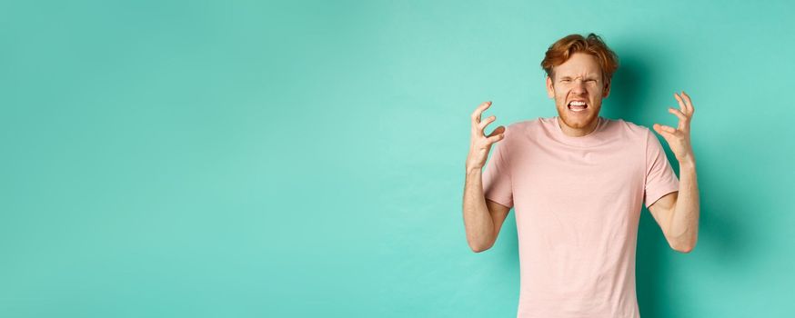 Annoyed and pissed-off redhead guy looking angry, shouting and grimacing, shaking hands aggressive, standing furious against mint background.